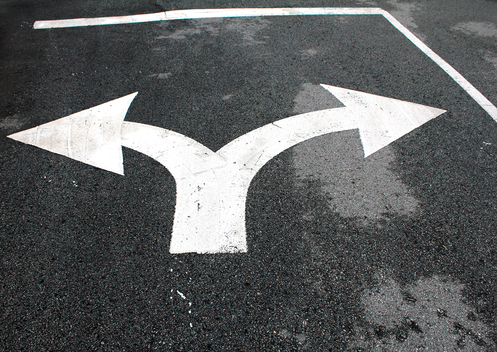 Weiße Straßenmarkierung mit nach links und rechts zeigenden Pfeilen auf dunklem Asphalt, die eine Richtungsänderung anzeigt.