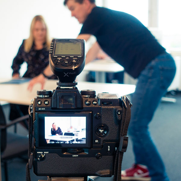 Eine Digitalkamera auf einem Stativ fängt eine Szene mit zwei Personen in einer Büroumgebung ein. Der Kamerabildschirm zeigt eine sitzende Person an einem Schreibtisch, während eine andere Person sich im Raum zu bewegen scheint.