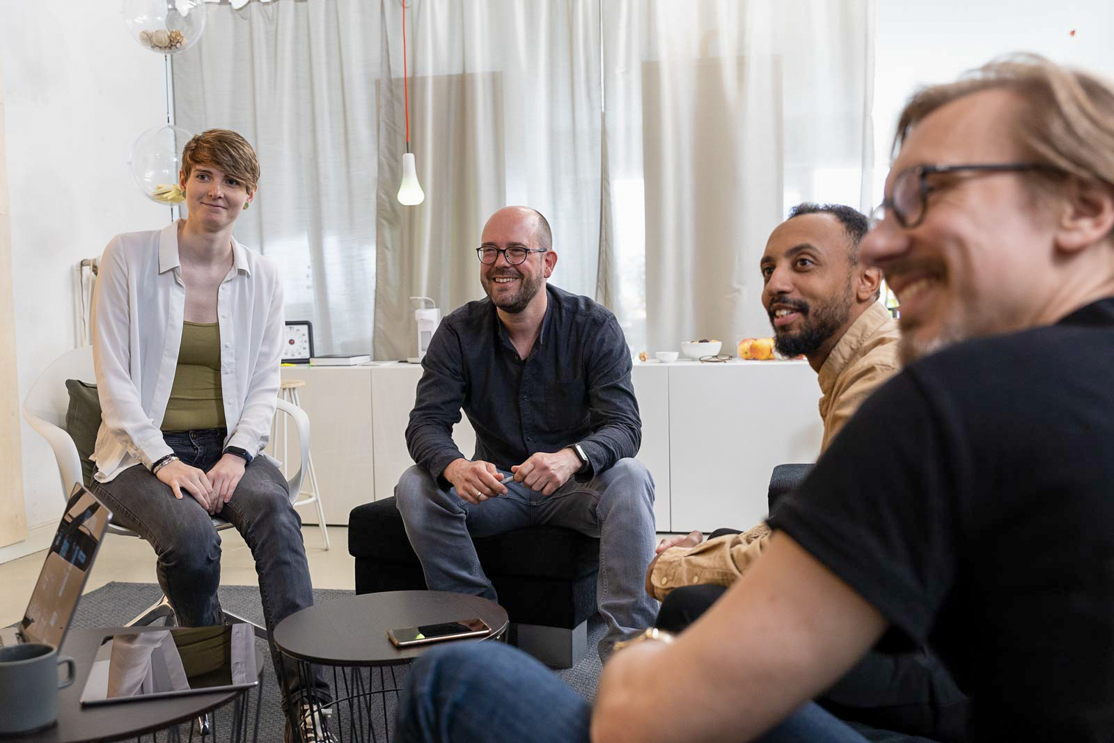 Eine Gruppe von vier Personen sitzt in einer ungezwungenen Büroumgebung und ist in eine lebhafte Diskussion vertieft. Sie sitzen an kleinen Tischen mit Geräten und Notizbüchern. Im Hintergrund sind transparente Vorhänge und minimalistisches Dekor zu sehen.