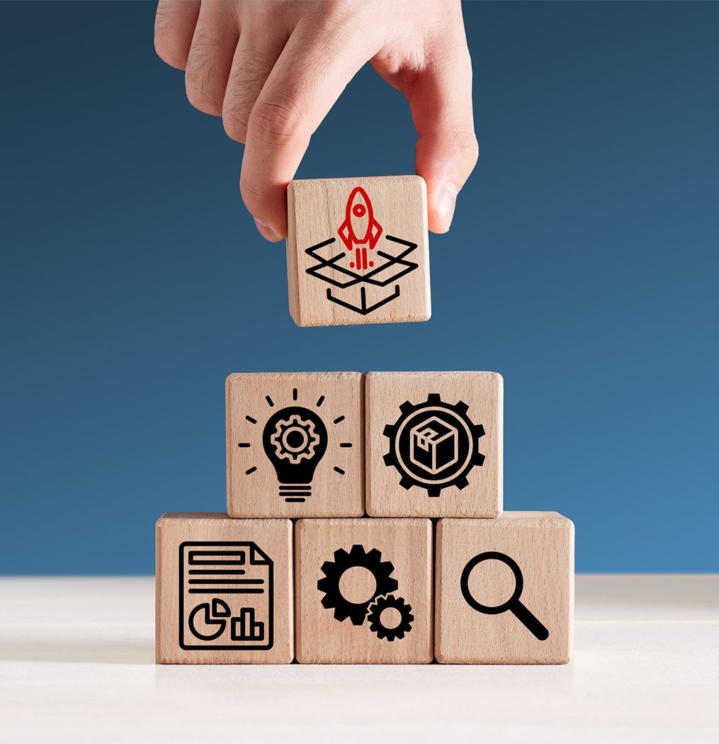Eine Hand stapelt Holzblöcke mit den Symbolen einer Rakete, einer Glühbirne, eines Zahnrads, einer Tabelle, eines Würfels und einer Lupe.